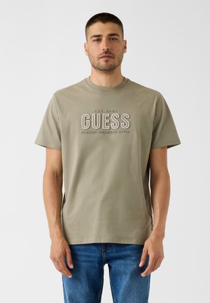 Man draagt beige "GUESS" T-shirt en blauwe spijkerbroek en staat voor een effen witte achtergrond.