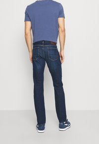 Dunkelblaue Jeans mit geradem Schnitt, Fünf-Taschen-Design und braunem Lederpatch am Bund, kombiniert mit einem grauen T-Shirt und blauen Sneakers.