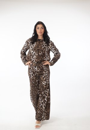 Woman standing with hands on hips wearing matching brown and black leopard print long-sleeve shirt and wide-leg pants, white heeled sandals.