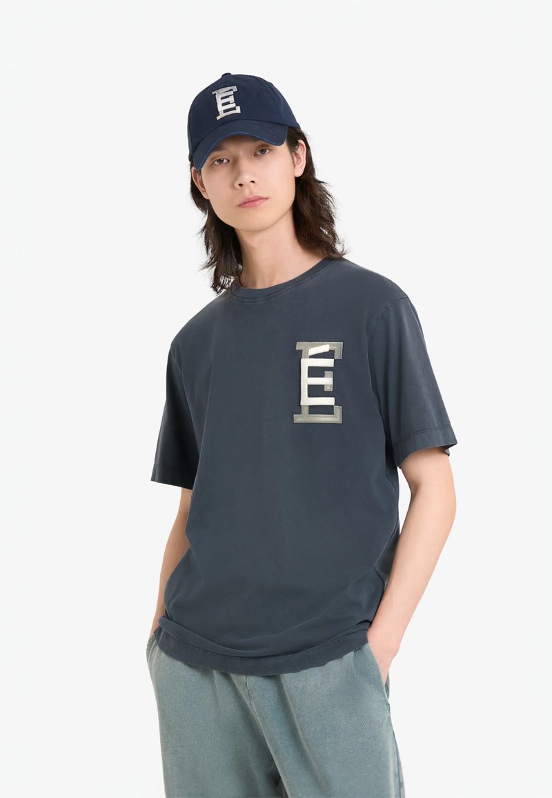 T-shirt en coton bleu foncé avec un logo métallique sur la poitrine gauche, associée à un pantalon vert clair légèrement texturé et une casquette assortie.