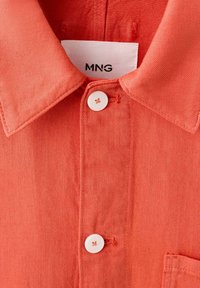 Chemise orange corail avec col classique, boutons blancs et étiquette de marque MNG visible à l'intérieur du col.