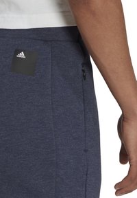 dunkelblaue Baumwoll-Sweatpants aus weichem Material mit einer seitlichen Reißverschlusstasche und einem Adidas-Logo-Patch auf der Rückseite.