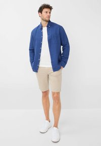 Camicia azzurra di lino con bottoni sopra una t-shirt bianca, pantaloni corti beige e sneakers bianche. La camicia presenta una tasca sul petto e maniche arrotolate.