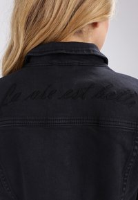 Giacca di jeans nera con la frase ricamata "La vie est belle" sul retro. Il colletto e le cuciture sono visibili, evidenziando il tessuto texturizzato.
