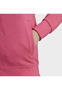 Mikina z fleesu v jasně růžové barvě, vybavená klokáním kapucí a žebrovanými manžetami. Hladká textura s jemnými švy.