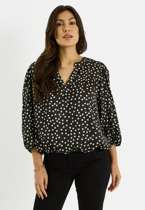 Mujer de cabello largo y oscuro que lleva una blusa negra con lunares blancos y pantalones negros, de pie frente a un fondo blanco liso.