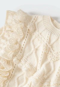 Krämfarbener gestrickter Pullover mit aufwendigen Zopfmuster. Akzentuiert mit zarter Spitzenbesatz und gewellten Kanten. Weiche Textur.