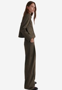 Olivgrüner, taillierter Blazer mit zwei Taschen und Knopfverschluss, kombiniert mit weiten Hosen aus passendem Stoff und in derselben Farbe.