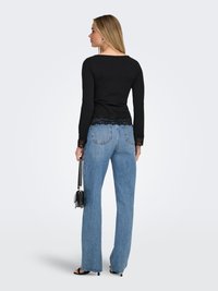 Vrouw met haar rug naar de camera, draagt een zwart shirt met lange mouwen en kanten rand, blauwe spijkerbroek, zwarte hakken, en houdt een kleine zwarte handtas naast zich vast.