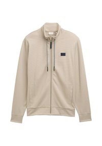 Giacca beige con zip e colletto alto, tessuto strutturato, tirazip nero e tasche laterali. Presenta un piccolo etichetta nera sul petto.