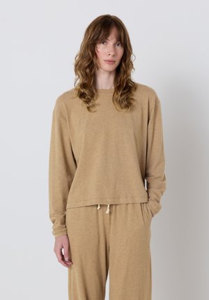 Nainen, jolla on laineikkaat ruskeat hiukset ja joka on pukeutunut löysään, pitkähihnaiseen beigeen yläosaan ja vastaaviin housuihin, joissa on nyöritys, seisoo valkoista seinää vasten.