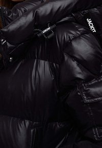 Czarna kurtka puchowa z błyszczącym, pikowanym wzorem, wyraźnym wypełnieniem, zamkiem błyskawicznym i ozdobnymi elementami z logo marki.