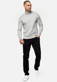 Man draagt een lichtgrijze coltrui, zwarte broek en witte sneakers, staat met één hand in de zak tegen een witte achtergrond.