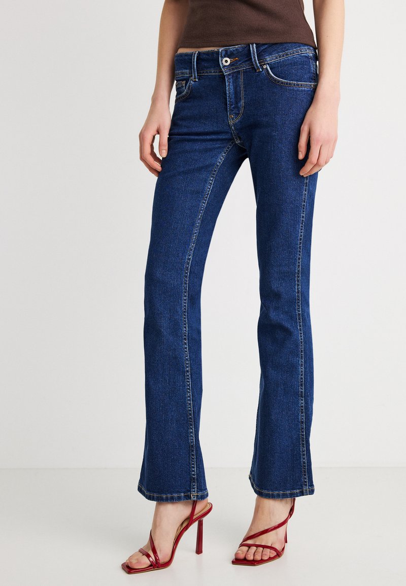 Blauwe denim flared jeans met een taille op middenhoogte, vijf zakken en contrasterende stiksels. Gecombineerd met rode sandalen met hoge hakken en bandjes.