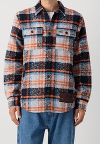 Kostkovaná flanelová košile v námořnické modré, oranžové a bílé barvě. Obsahuje dvě náprsní kapsy, zapínání na knoflíky a označení značky na boku. Texturovaná tkanina.