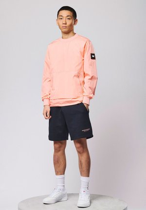 Junger Mann, der eine hellrosa Windjacke, schwarze Shorts, weiße Socken und weiße Turnschuhe trägt, vor einem einfarbigen Hintergrund steht.