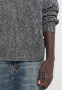 Άτομο που φοράει γκρι πλεκτό πουλόβερ με τα μανίκια σηκωμένα και ξεθωριασμένο μπλε τζιν, με το δεξί χέρι να κρέμεται χαλαρά δίπλα του.