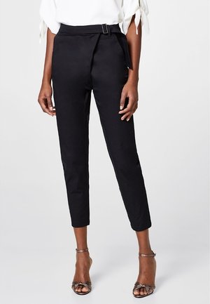 Zwarte hooggerokte taps toelopende broek met stoffen riem, gedragen met metallic open teen enkelriem hakken en een witte top met gestrikte mouwen.