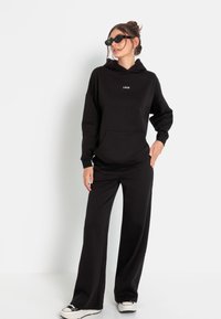 Schwarzer Hoodie mit Fronttasche und Logo; kombiniert mit schwarzen weiten Hosen und weißen Sneakers; Modell trägt Sonnenbrillen und posiert vor einem einfarbigen Hintergrund.