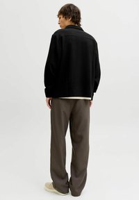 Czarna, teksturowana kurtka-koszula z kołnierzem, zestawiona z luźnymi, brązowymi spodniami. Szare tło. Prosty wzór; brak widocznych guzików i wzorów.