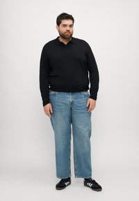 Polo shirt nero a maniche lunghe, jeans azzurri a gamba dritta, sneaker nere con strisce bianche, in piedi contro uno sfondo bianco semplice.
