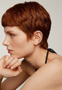 Pelo corto, rojo y texturizado, peinado cerca de la cabeza. El sujeto lleva un pendiente de aro plateado y reposa una mano bajo la barbilla.
