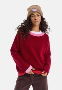 Czerwony sweter z różowym prążkowanym dekoltem i mankietami; model nosi pod spodem jasną koszulę. Beżowa dzianinowa czapka z fioletową metką.