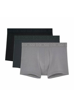 Hemdloser Mann, barfuß stehend, trägt graue Boxershorts mit "HOM" Logo am Bund vor einem schlichten hellen Hintergrund.