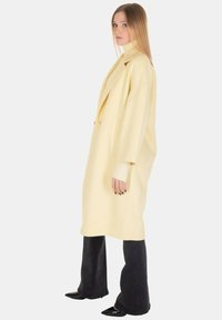 Lungo cappotto giallo con forma strutturata, grandi risvolti e tasche laterali. Indossato sopra un dolcevita aderente e abbinato a pantaloni neri svasati.