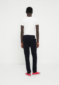 Ναυτικό μπλε παντελόνι φόρμας με πλαϊνές τσέπες και στενή εφαρμογή, συνδυασμένο με ένα απλό λευκό μπλουζάκι και κόκκινα αθλητικά παπούτσια σε ουδέτερο φόντο.