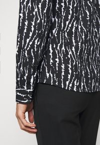 Camicia a righe in bianco e nero con un design seghettato, maniche lunghe, chiusure a bottoni e un orlo curvo, abbinata a pantaloni scuri.