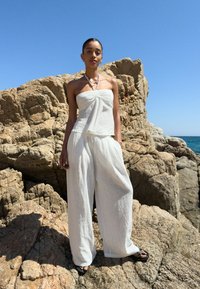 Bijela lanena top bez naramenica s čvor detaljem, uparen s opuštenim bijelim lanenim hlačama. Crne ravne sandale. Stjenovita plaža u pozadini i vedro nebo.