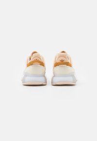 Sneakers with a soft peach mesh upper, white sole, and yellow accents. Features a textured heel and three-stripe branding at the back.
