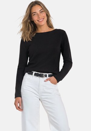 Femme souriante aux cheveux mi-longs portant un haut noir à manches longues, un pantalon blanc et une ceinture noire, une main dans la poche.