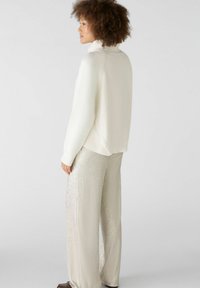 Weißer gerippter Pullover mit hohem Kragen, kombiniert mit glänzenden, silbernen, weit geschnittenen Hosen, seitlich vor einem neutralen Hintergrund gezeigt.