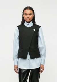 Čierna bezrukávová vesta s asymetrickým predným dizajnom, obsahujúca dve predné vrecká a malé biele logo, nosená cez svetlomodrú košeľu na gombíky.