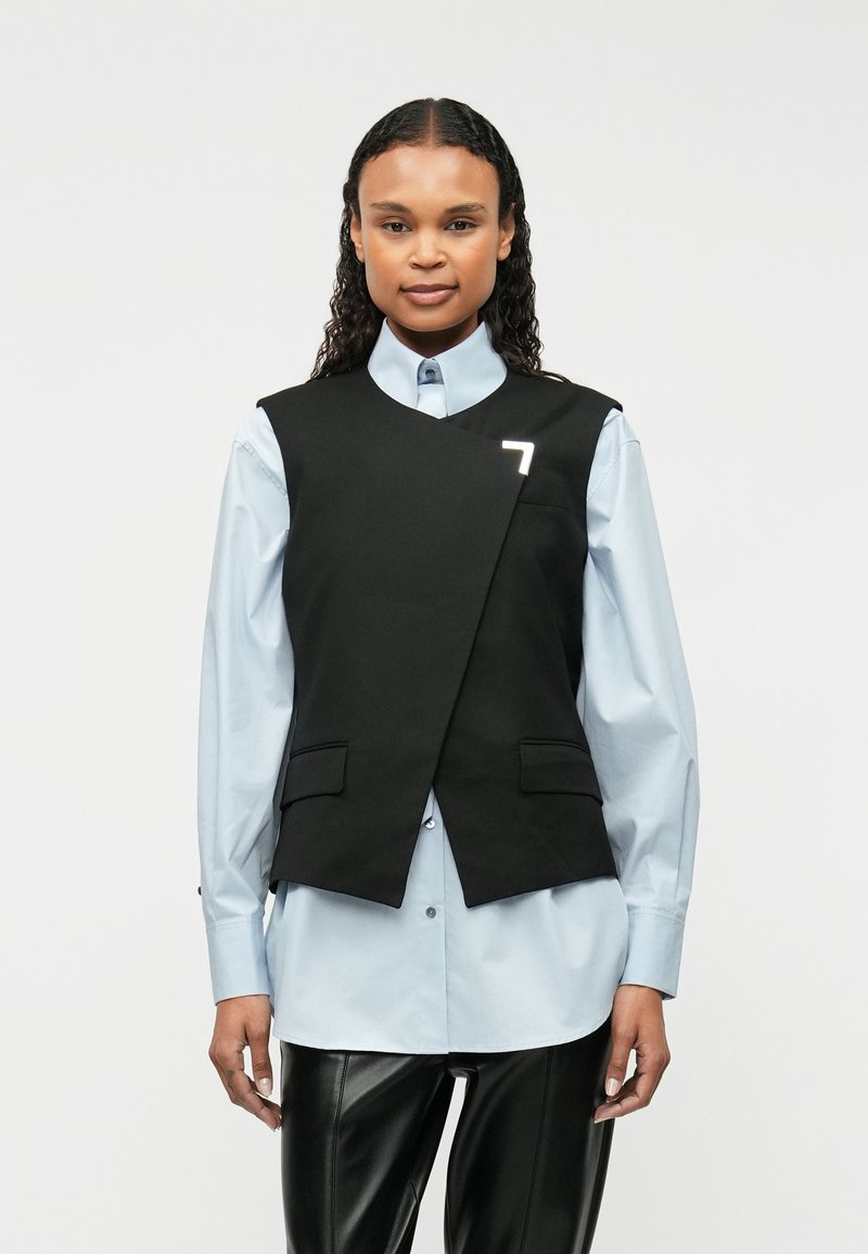 Čierna bezrukávová vesta s asymetrickým predným dizajnom, obsahujúca dve predné vrecká a malé biele logo, nosená cez svetlomodrú košeľu na gombíky.