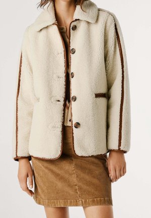 Kobieta ubrana w kremową kurtkę sherpa z brązowym wykończeniem i guzikami, w połączeniu z brązową spódnicą sztruksową.