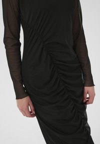 Black dress with long sheer sleeves, featuring diagonal gathering and ruching on the front. Smooth texture with a fitted design.