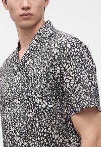 Homme portant une chemise à manches courtes noire et blanche à motifs avec une poche avant, montré de la poitrine vers le haut sur un fond uni.