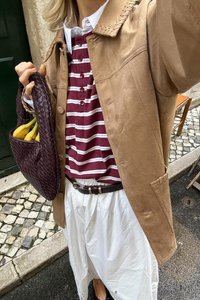 Beige Wildlederjacke mit großem Kragen, bordeaux-rot-weiß gestreiftes Hemd mit Knöpfen, weiße Skirt, braune geflochtene Tasche mit Bananen.