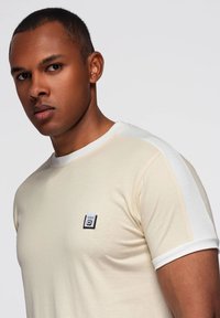 Béžové tričko s krátkým rukávem a bílými akcenty, kulatým výstřihem a malou etiketou na přední straně. Měkká látka s hladkou strukturou.
