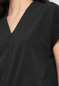 Haut noir à manches courtes avec un décolleté en V profond, une texture lisse et une coupe décontractée, mettant en valeur des lignes épurées et un design minimaliste.