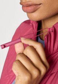 Nike vetrovka s vysokým golierom, hlboká ružová farba, so zipsom a nastaviteľnou šnúrkou, držaná rukou. Hladká textura.