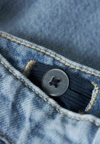 Nærbilde av en mørk knapp festet på lyseblå denimstoff med synlige sømmer og en knapphull.