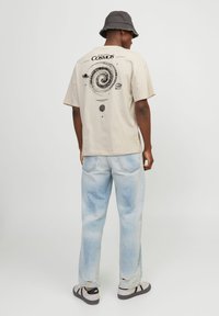 T-shirt en coton beige avec un graphique de galaxie en spirale noir au dos ; associé à un jean en denim délavé et des baskets noir et blanc.