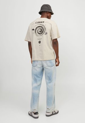 Beige Baumwoll-T-Shirt mit spiralgalaxie Grafik in Schwarz auf dem Rücken; kombiniert mit hellwaschblauen Jeans und schwarz-weißen Sneakern.