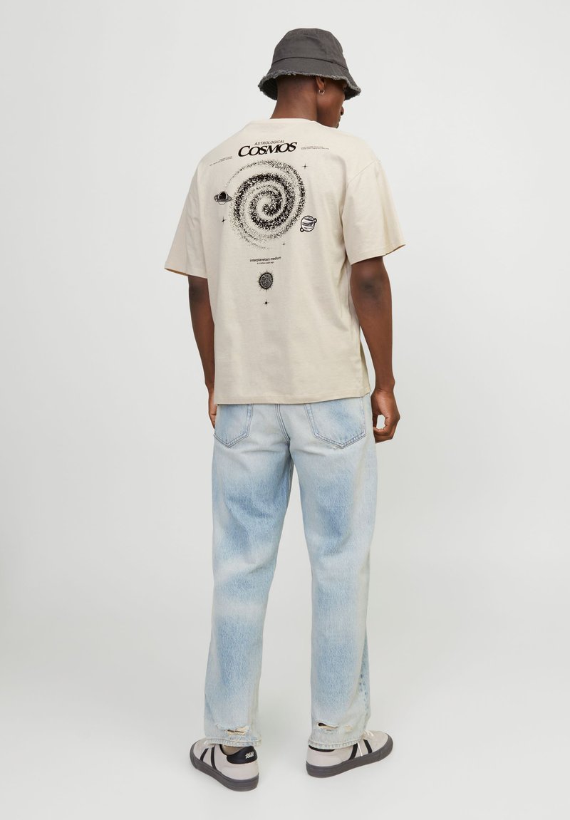 T-shirt en coton beige avec un graphique de galaxie en spirale noir au dos ; associé à un jean en denim délavé et des baskets noir et blanc.