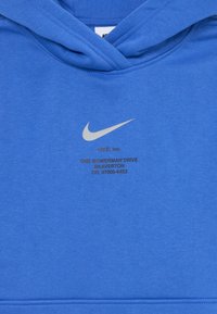 Modra kapuca iz mehkega materiala, z sprednjim žepom, kapuco s vrvico in sivo Nike logo na prsih s besedilom pod njim.