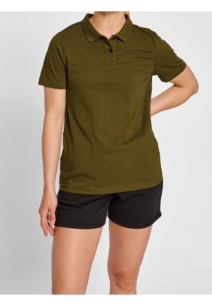 Persoon draagt een olijfgroen poloshirt en zwarte korte broek, staand met één hand op de heup en de andere ontspannen langs het lichaam.